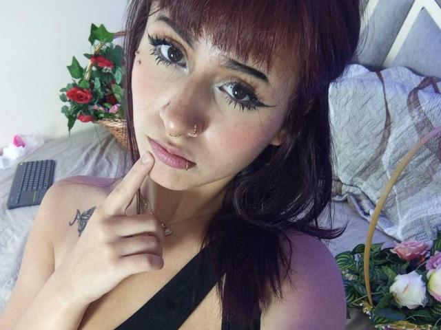 Close-up de KatheLux, uma mulher de pele clara com cabelo castanho escuro, delineador preto intenso e um piercing no nariz. Ela está tocando o lábio com o dedo, usando uma alça preta. Arranjo floral ao fundo.