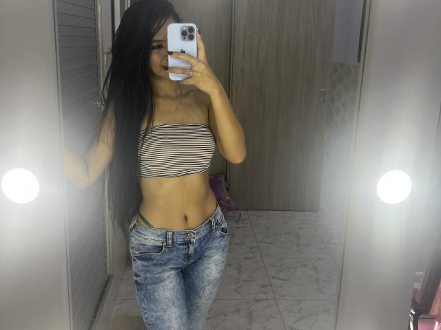 EstrellaXHot, una atractiva mujer latina con cabello largo y negro, se toma un selfie frente al espejo. Ella lleva una blusa negra y blanca a rayas, sin tirantes, y unos jeans azules ajustados. Su mano cubre su teléfono, del tamaño de su pene..