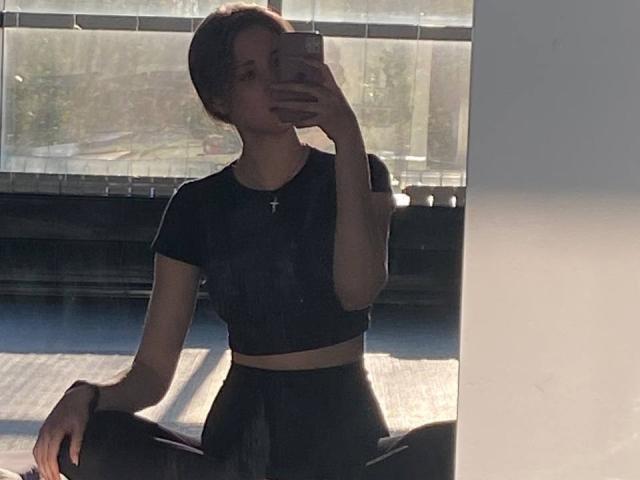 Fotografia in silhouette di KenaCly, con capelli castani corti, che tiene il telefono vicino al viso, indossa un top corto nero e pantaloni a vita alta. pantaloni, seduto in una finestra illuminata dal sole, in ombra, teso, con una mano sul ginocchio, una croce al collo..