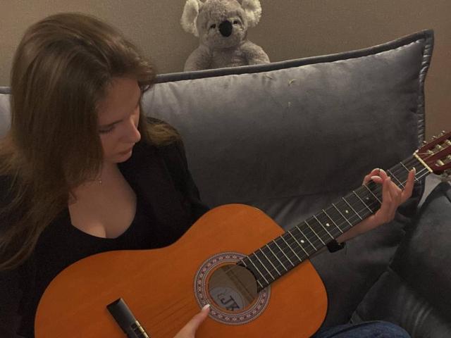 KenaCly, con el pelo largo y castaño, vestida con una blusa negra, rasguea una guitarra acústica en un sofá oscuro, con un koala de peluche. Detrás de ella, un cuchillo negro en su mano derecha.