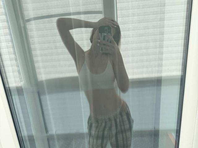 Autoportrait miroir de KenaCly, une main sur la tête, portant un soutien-gorge blanc et un pantalon à carreaux, reflétant la lumière vive d'une fenêtre. Appareil photo à la main, peau claire, pose décontractée, légère courbure du corps..