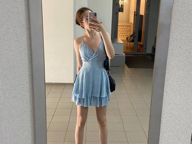 KenaCly prend un selfie devant un miroir dans un espace intérieur lumineux, vêtue d'une robe bleu clair à décolleté en V et à plusieurs niveaux qui met en valeur sa silhouette. Silhouette mince et petites seins. Tenant un sac à main noir. Reflète une pose confiante..