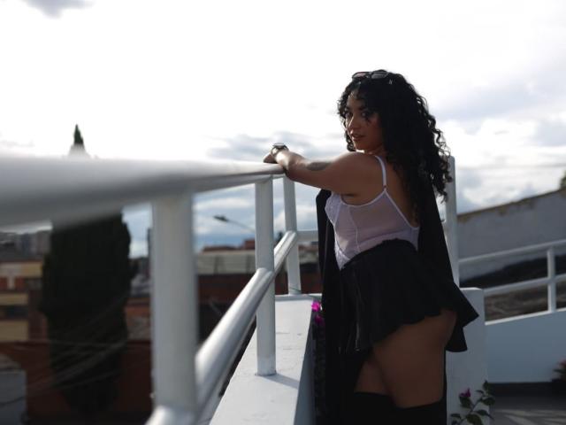 Fotografia de CarmilleZlata, uma mulher curvilínea com longos cabelos cacheados pretos, apoiada em um corrimão branco. Ela veste uma blusa branca transparente e lingerie preta, exibindo suas nádegas grandes. Luz solar intensa.