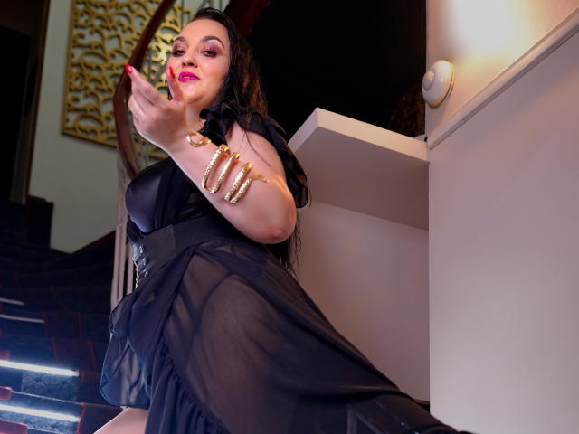 KattSuunny with big ass, black dress, and long black hair, wearing gold bracelets, pink lipstick, and looking down, standing on stairs, holding a phone in her right hand.
