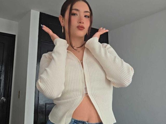 Stardustt with light skin and dark hair, wearing an open white ribbed shirt revealing a small cleavage and midriff, hands up, pouty expression, standing in front of a black door.