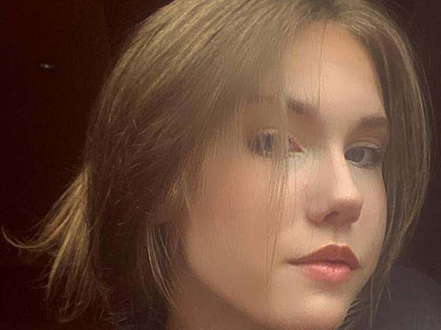 Gros plan de KenaCly, une jeune femme aux cheveux bruns clairs, légèrement en désordre, et à la peau pâle. Elle regarde directement l'appareil photo avec une expression neutre, un léger rougissement sur ses lèvres..