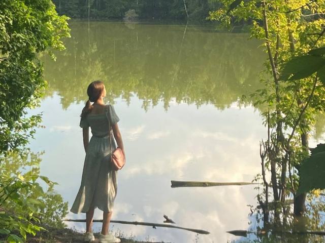 KenaCly está de pie cerca de un lago tranquilo, vestida con un vestido gris y sosteniendo una bolsa marrón. La luz del sol se filtra a través de los árboles verdes, proyectando sombras sobre el agua. Ella mira a lo lejos..