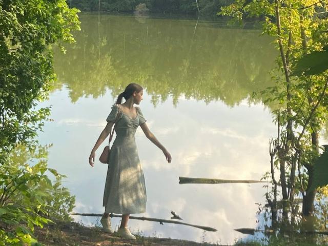 KenaCly, gekleed in een groene jurk, loopt langs een door de zon verlichte oever, omringd door dichte begroeiing. De reflecties dansen op het water. Ze draagt een bruine schoudertas, die een schaduw werpt. Heel rustgevend..