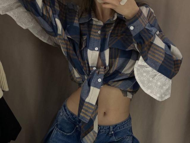Fotografía de KenaCly con una camisa de cuadros anudada, dejando al descubierto su abdomen, y con jeans azules. Su mano está levantada cerca de su rostro, creando una pose ligeramente provocativa. Ombligo visible. Casual, ligeramente sexy..