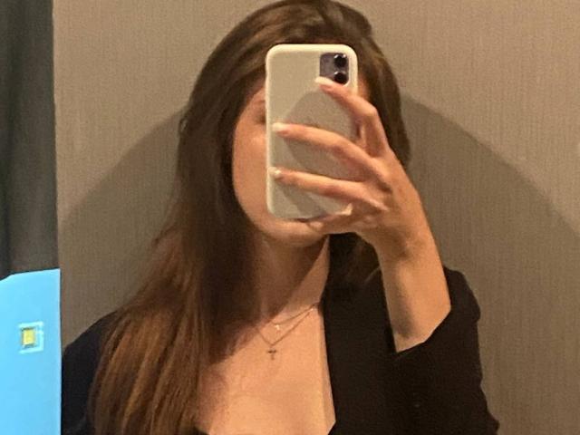 KenaCly se toma un selfie frente a un espejo, sosteniendo un iPhone blanco frente a su cara, lo que la oculta parcialmente. Ella tiene el pelo largo y castaño, lleva una blusa negra con un collar, y muestra un ligero escote..