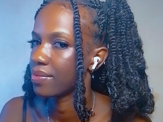 Close-up of StylishAurora, an attractive Black woman with shiny, oiled skin and long, thick, braided hair. She wears white earbuds and a necklace, gazing seductively at the camera. Blue light highlights her face.