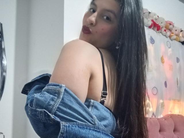 MadissonFontaine, a young woman with long black hair, gazes over her shoulder, wearing a denim jacket off her shoulders, revealing black lingerie. Her face has a slight blush, sexy expression.