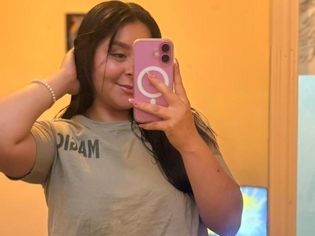 EmaJames with long black hair, smiling, holding a pink phone with a white circular design, wearing a grey "PIRAM" t-shirt, looking at the mirror, sexy, casual selfie, wrist bracelet.