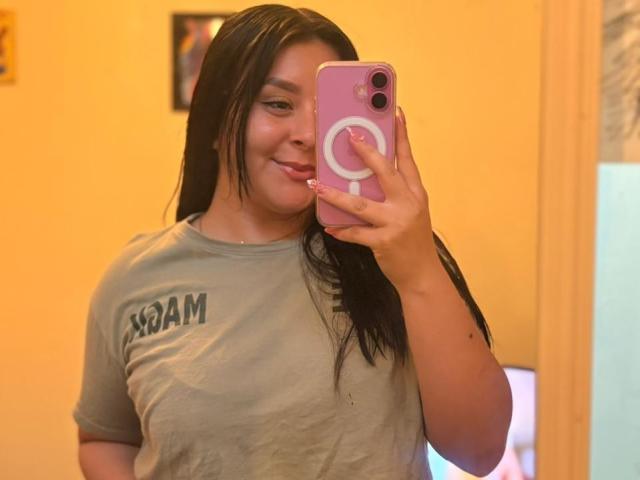 EmaJames takes a selfie in a yellow-walled room, holding a pink phone with a white ring. She's smiling, wearing a wet grey "BOOM" t-shirt, showing her slightly large breasts.