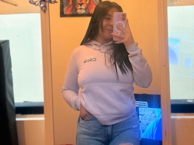 EmaJames steht vor einem Spiegel und macht ein Selfie mit ihrem rosa Handy. Sie trägt eine weiße Kapuzenjacke mit dem Aufdruck "ASOS" und helle blaue Jeans. Ihre Hand ist in ihrer Tasche..
