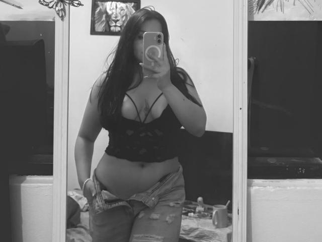 Black and white mirror selfie of EmaJames, showing her curvy figure with large breasts in a low-cut lace top and unbuttoned, ripped jeans. Her phone covers her face. Bedroom background.
