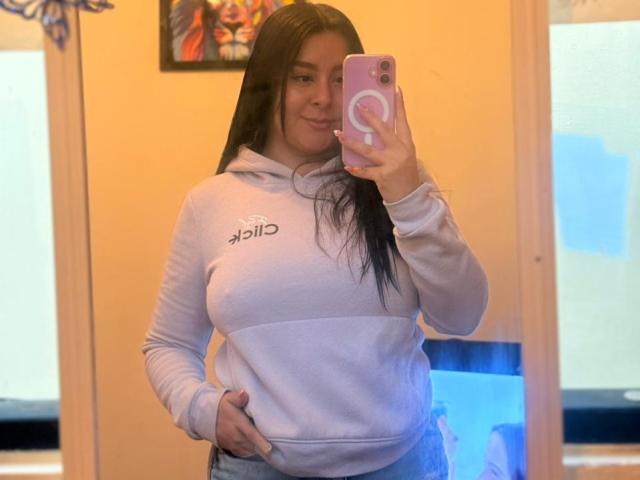 EmaJames with long black hair, wearing a white Asics hoodie, taking a mirror selfie with a pink phone. She has a hand in her hoodie pocket, looking confident. Blue and orange background.