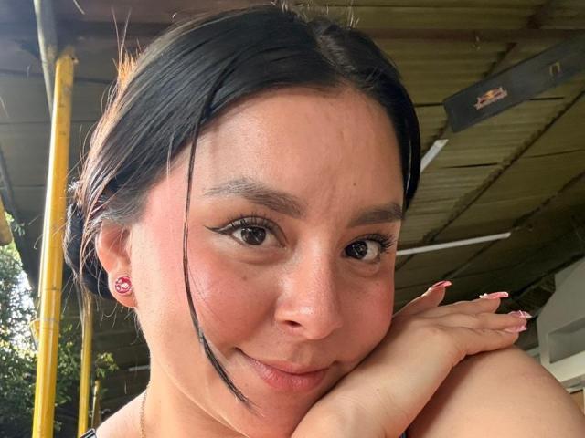 Close-up of EmaJames, a woman with dark hair, fair skin, and black eyeliner, resting her chin on her hand, smiling subtly. Yellow beams and roof in background. Her nails are painted pink.
