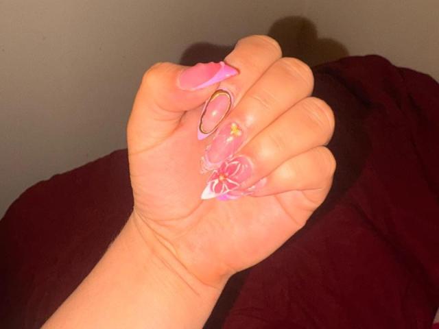 Close-up of EmaJames' hand with pink nail polish, holding a ring on a black background. Fingers spread, showcasing nails and ring. Bright light highlights hand, creating a stark contrast.