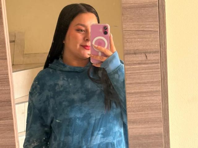 EmaJames, a woman with long black hair, takes a mirror selfie with a pink phone. She wears a blue tie-dye hoodie, holding the phone in her right hand. Fucks up her face slightly.