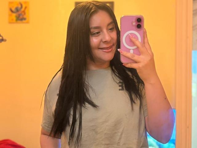 EmaJames, with long black hair, smiles while taking a selfie with a pink phone. She's wearing a gray T-shirt, her hand gripping the phone, light orange background. Fucking sexy.