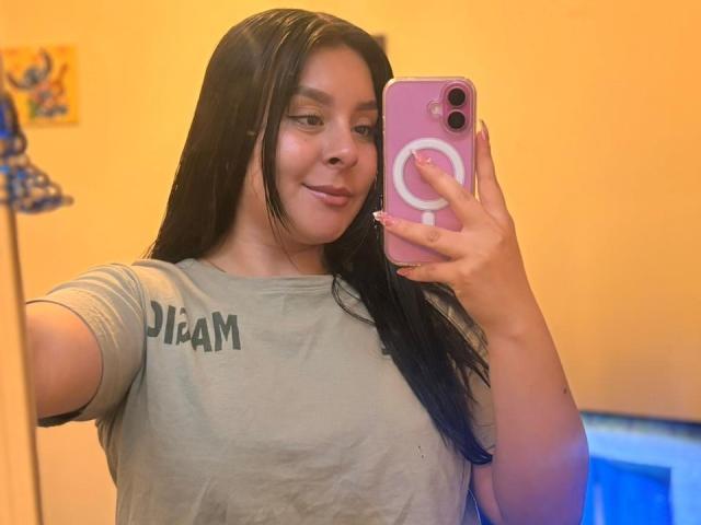 EmaJames takes a selfie with a pink phone, holding it in her right hand. She has long black hair, wears a gray "DIAM" t-shirt, and smiles subtly. Background is orange and blue.