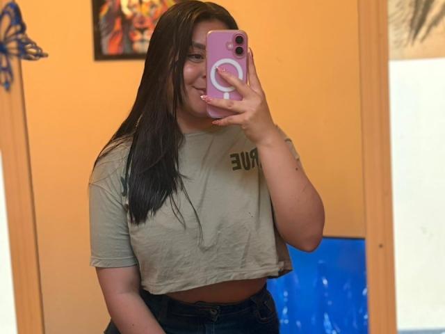 EmaJames takes a mirror selfie, holding a pink phone with a white ring. She's wearing a wet, tight, beige crop top, showing her small breasts. Long black hair, sexual tension.