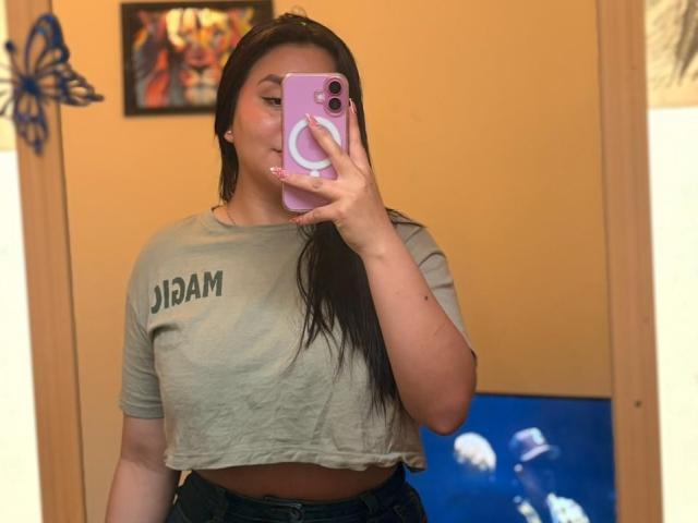 EmaJames with long black hair takes a selfie in a mirror, holding a pink phone with a glowing ring. She's wearing a cropped gray "IBM" t-shirt, showing her midriff. Blue wall and butterfly decoration in background.