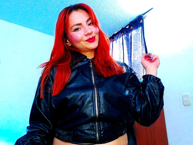EmelySofia with vibrant red hair, smiling, wearing a black leather jacket, holding a hair tie, blue and white background, bright colors, confident pose, slight shadow on the right.