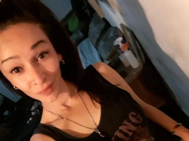Foto em close-up de LunaCreciente, uma mulher de pele clara, cabelos escuros e seios pequenos, vestindo uma camisa preta. colar, e brincos de argola, sentada em um quarto bagunçado, rosto sombreado, olhar sedutor..