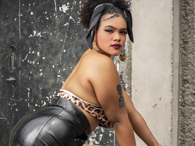 Image numérique d'AliceJelanni, avec une peau foncée, des cheveux bouclés attachés dans un bandana noir, un haut à imprimé léopard et un pantalon en cuir noir. Elle se penche en avant, jetant un coup d'œil en arrière, et montre un tatouage sur son bras droit. Tension sexuelle.