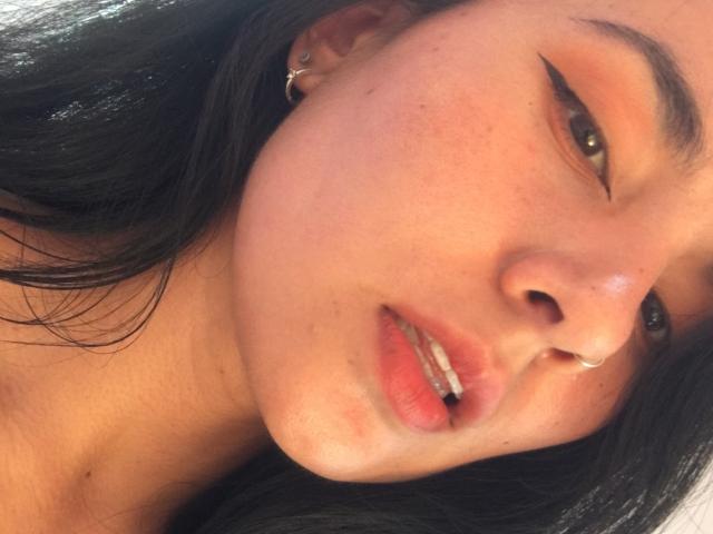 Primer plano del rostro de Natii69, con cabello negro, piel clara y un piercing en la nariz. Ella tiene ojos marrones, labios ligeramente entreabiertos que muestran los dientes, y un pequeño arete de oro. Mirada increíblemente sensual e intensa..