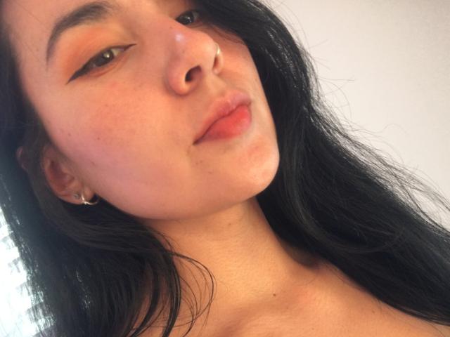 Close-up photo of Natii69 with long black hair, fair skin, and a nose piercing. She gazes slightly to the side with a subtle, sultry expression, lips slightly parted.