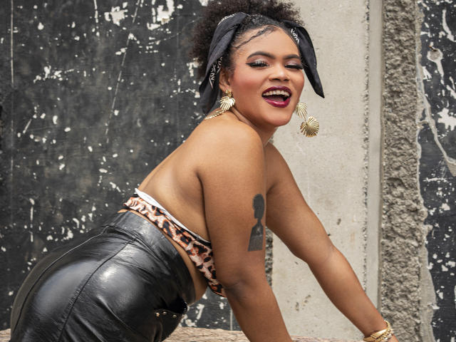 AliceJelanni leans forward on a concrete ledge, wearing a leopard-print strapless top and black leather pants. She has dark skin, red lipstick, large gold earrings, and a key tattoo on her arm. Her expression is flirtatious.