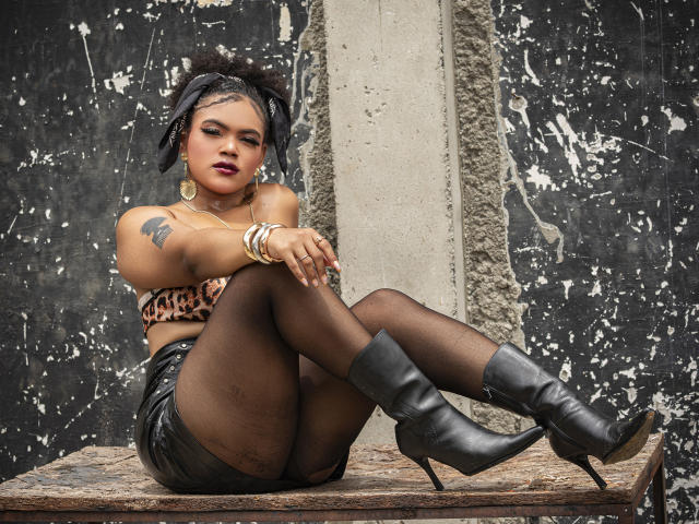 AliceJelanni, donkerhuidig, met zwart haar, zit op een ruwe rand, met gekruiste benen, en draagt een beha met luipaardprint, zwart korte broek, netpanty's en zwarte laarzen. Tatoeage op de linker schouder. Verdomd sexy..