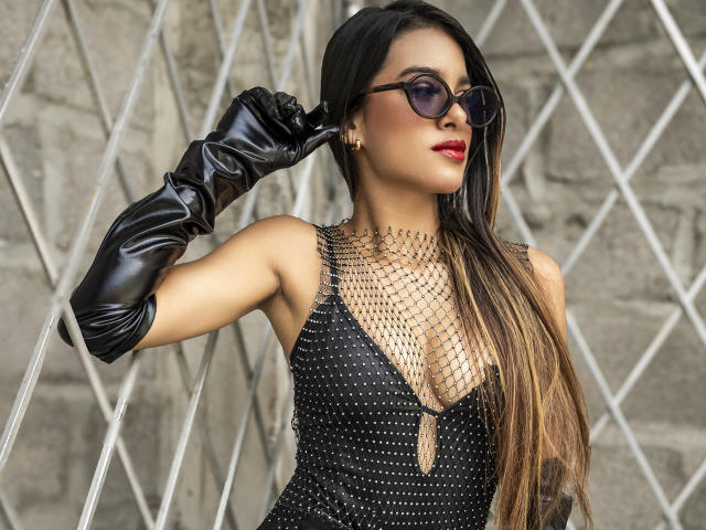 Vanelope Russo se tient devant un mur à motifs losanges, vêtue d'une robe noire ornée de clous, de gants noirs et de lunettes de soleil foncées. Elle ajuste ses cheveux, adoptant une pose confiante et séduisante avec du rouge à lèvres..