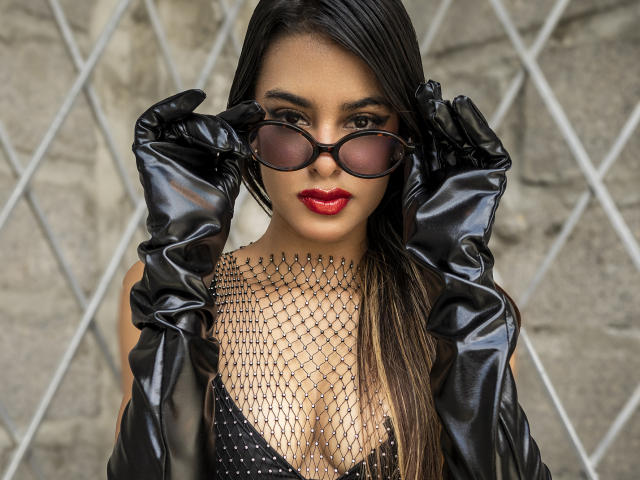Vanelope Russo, mit schwarzen Lederhandschuhen, die dunkle Sonnenbrille justierend, rotem Lippenstift, einem schwarzen Netzoberteil und Leder. Jacke, vor einem Zaun mit Rautenmuster. Leichte Ausschnitt, sexy, selbstbewusst, intensiver Blick.