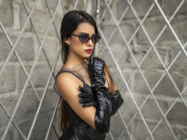 VanelopeRusso stands in front of a diamond-patterned wire fence, wearing black leather gloves, a sparkly black dress, and dark sunglasses. She has long black hair, red lipstick, and a sultry expression.