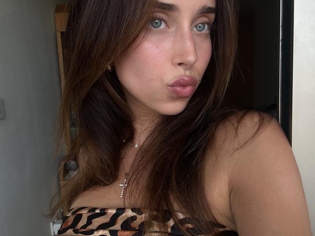 Primer plano de AdeleVibe con ojos azules, labios carnosos y cabello largo castaño. Llevaba una blusa con estampado de leopardo, y empujaba ligeramente sus grandes senos hacia adentro. La piel es clara con un ligero rubor..