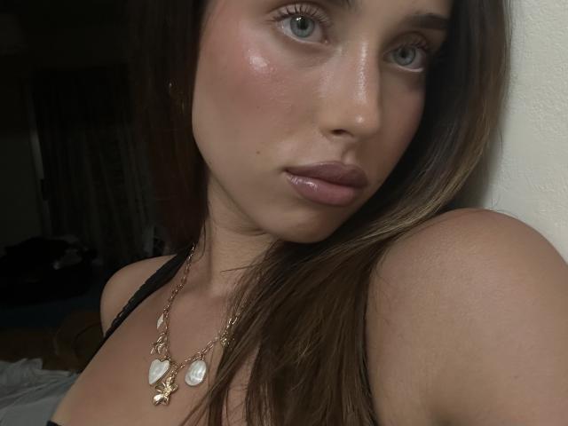 Close-up photo of AdeleVibe, a brunette woman with blue eyes, glossy pink lips, and a slight blush. She wears a gold necklace with heart-shaped pendants. Her shoulder is exposed. Fucking sexy.