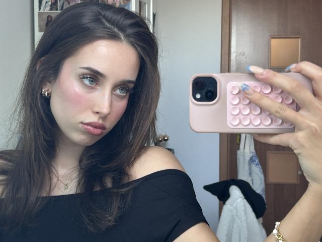 Un selfie de AdeleVibe con cabello castaño largo, ojos azules y rubor rosado. Ella sostiene un teléfono rosa con una funda de lunares y lleva puesto un top negro con un hombro descubierto. El fondo incluye una puerta y un perro blanco y negro..