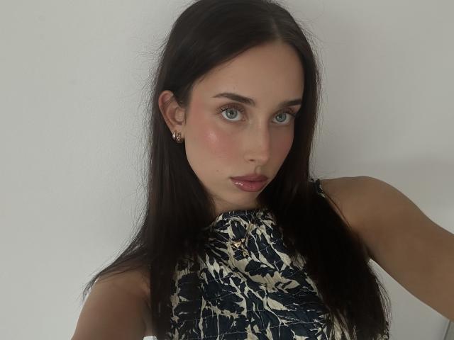 Adele, com seus longos cabelos pretos e lisos, olhos azuis e bochechas rosadas, veste um vestido preto e branco com padrão geométrico, sem mangas. topo. Ela está olhando diretamente para a câmera com a boca ligeiramente aberta. A tensão sexual é evidente..