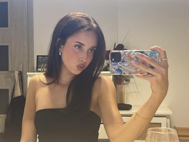 AdeleVibe, una donna castana con capelli lisci, si scatta un selfie davanti a uno specchio. Indossa un top nero senza spalline, con le labbra tumefatte e gli occhi azzurri. Il suo telefono ha una custodia colorata. La sua mano si trova sul suo pene..