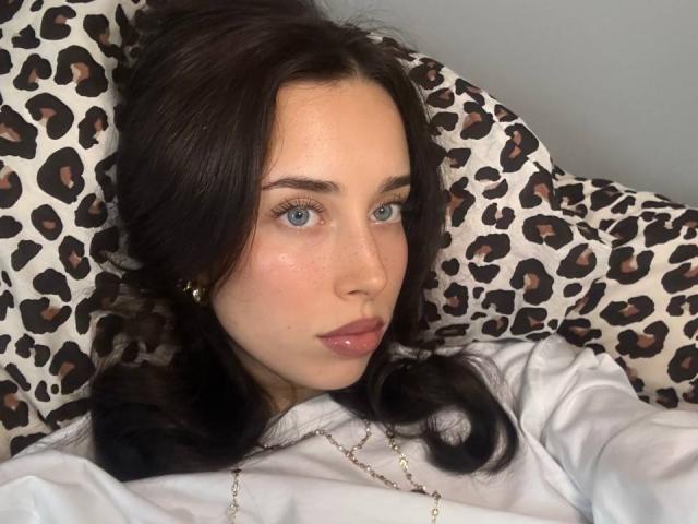 AdeleVibe, con piel pálida y ojos azules, está acostada sobre un cojín de estampado de leopardo, vistiendo una camisa blanca, con cabello negro ondulado y collar de oro. Increíblemente atractivo, con unos ojos azules que miran hacia adelante..