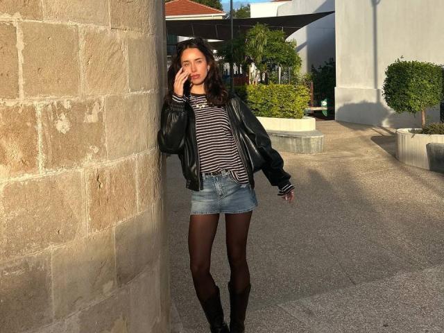 AdeleVibe se apoya contra una pared de piedra, vistiendo una chaqueta de cuero negra, una camisa a rayas, una falda vaquera y mallas negras. Ella está hablando por teléfono, proyectando una sombra en una calle soleada..