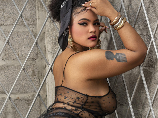 Alice Jelanni, à la peau foncée, aux formes généreuses, portant de la lingerie noire transparente, un foulard noir sur la tête, des boucles d'oreilles dorées, des bracelets, tatouage sur le bras supérieur. Se penche contre un mur à motif losange, la main sur la tête, expression sensuelle..