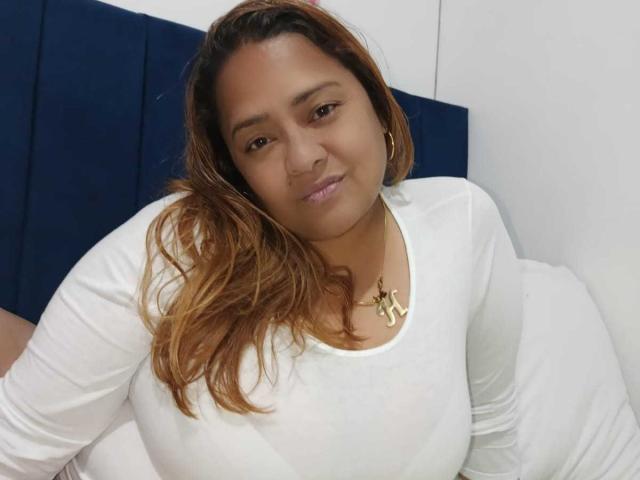 Fotografia de Monzerrat Boyer, com pele morena clara e cabelos castanhos longos e ondulados, vestindo uma blusa branca justa que realça seios grandes. Ela está sorrindo levemente, usando um colar de ouro. Há uma tensão sexual no ar..