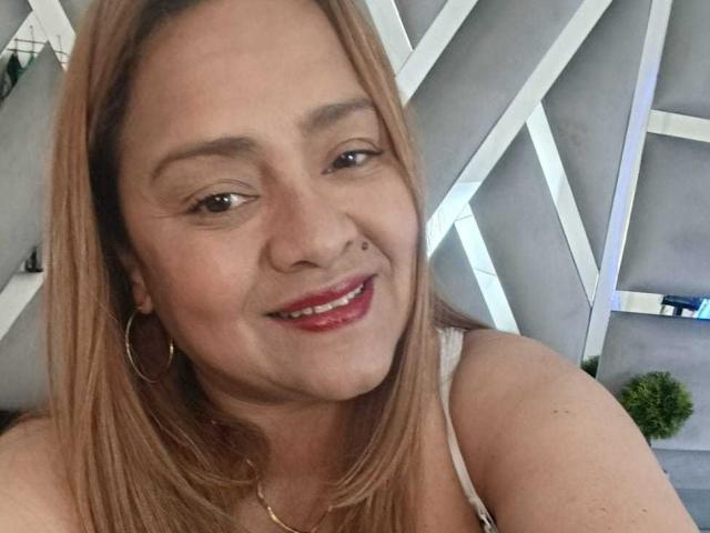 Close-up de Monzerrat Boyer com cabelo castanho claro, sorrindo com batom vermelho, vestindo uma regata branca e usando brincos de argola. e um colar de ouro. O fundo apresenta luzes geométricas brancas e plantas verdes..