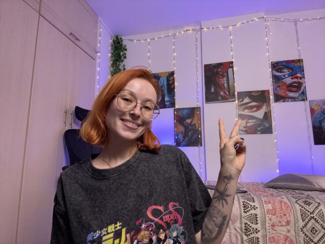 Sarah Rousen mit roten Haaren und Brille, die ein schwarzes T-Shirt mit einem Aufdruck trägt und mit ihrer tätowierten Hand den Mittelfinger zeigt. Hintergrund: Lichterketten, farbenfrohe Poster und ein Bett mit einer Decke mit Muster..