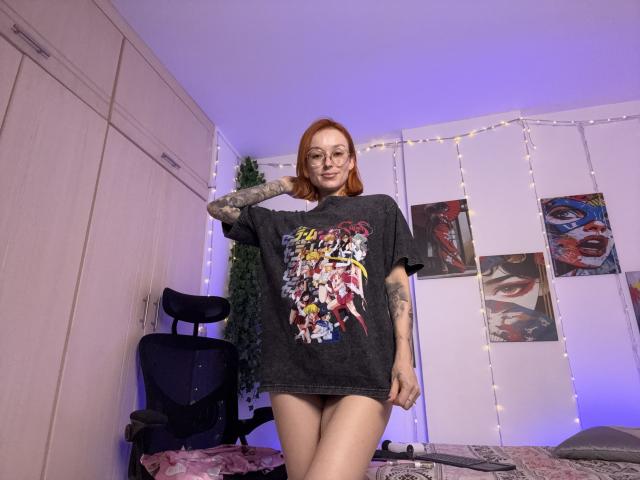 SarahRousen, a red-haired woman with glasses, stands in a room wearing a black graphic T-shirt, no pants, displaying tattoos. String lights, posters, and a chair in the background. Fucking sexy.
